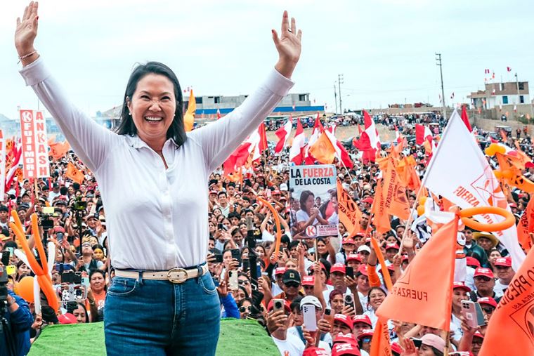 Lideresa política levanta los brazos ante una multitud con banderas peruanas y símbolos de campaña durante un mitin.